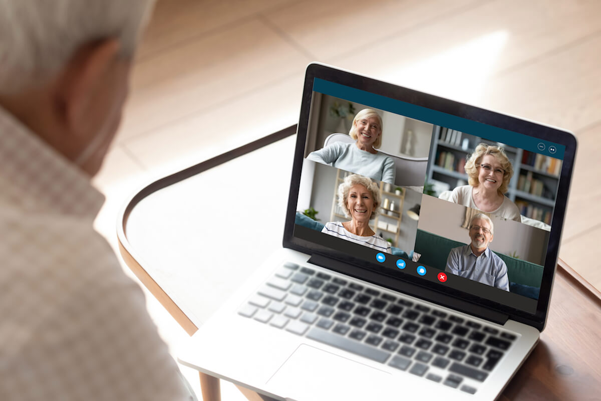 Old man makes videocall talking with relatives or friends by video conference app, pc screen view over male shoulder