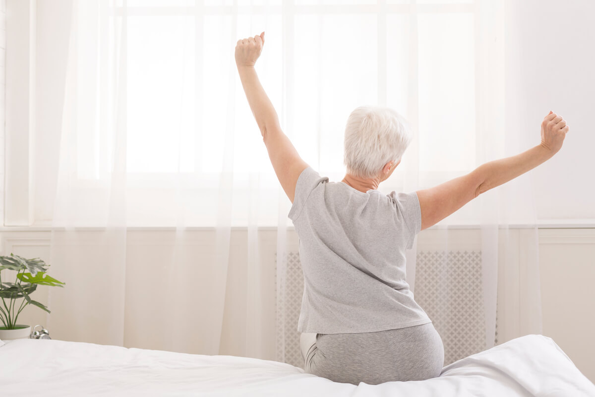 Senior woman sitting on her bed in morning, stretching with arms raised, back view, free space